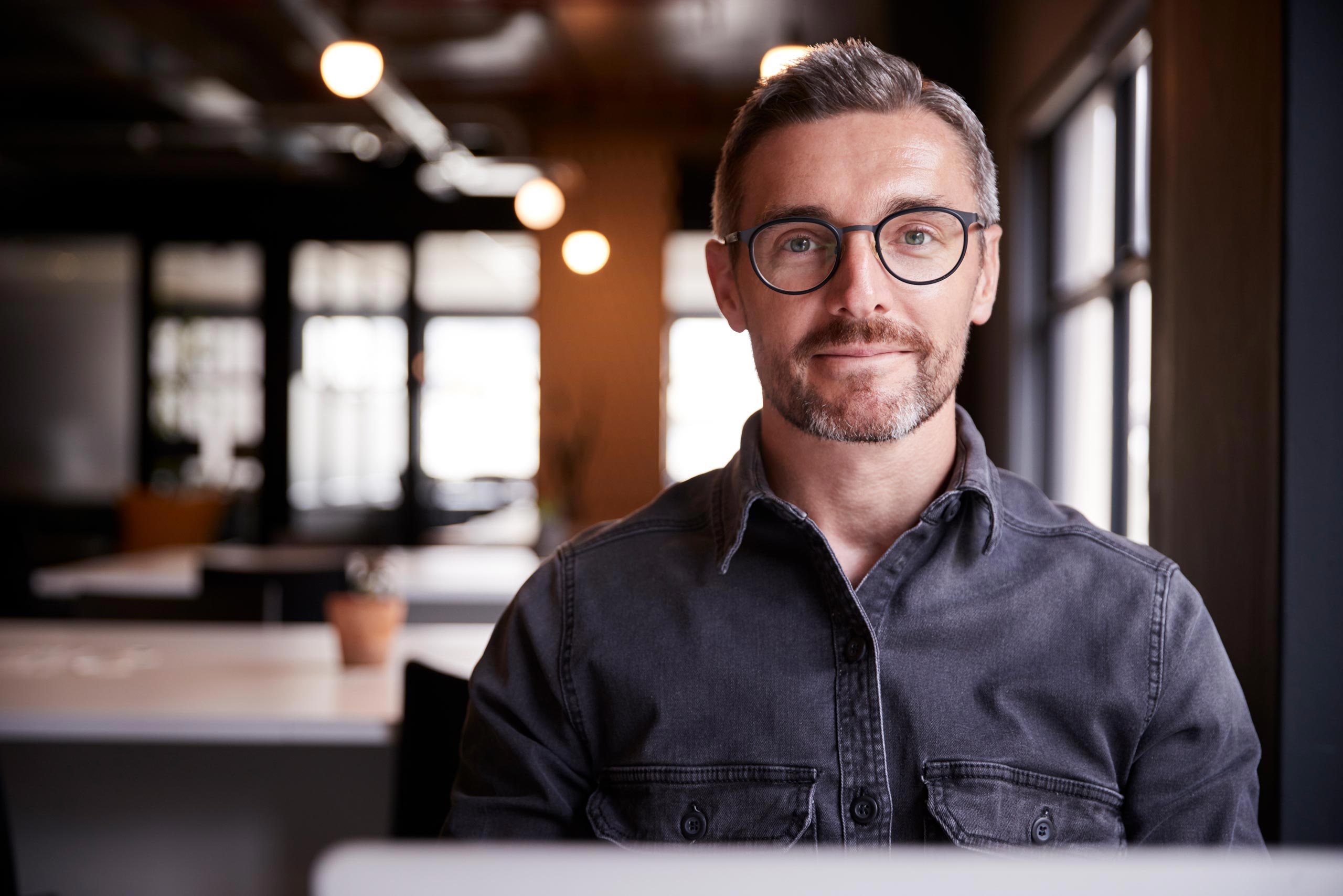 Smiling man wearing glasses in an open plan office space.