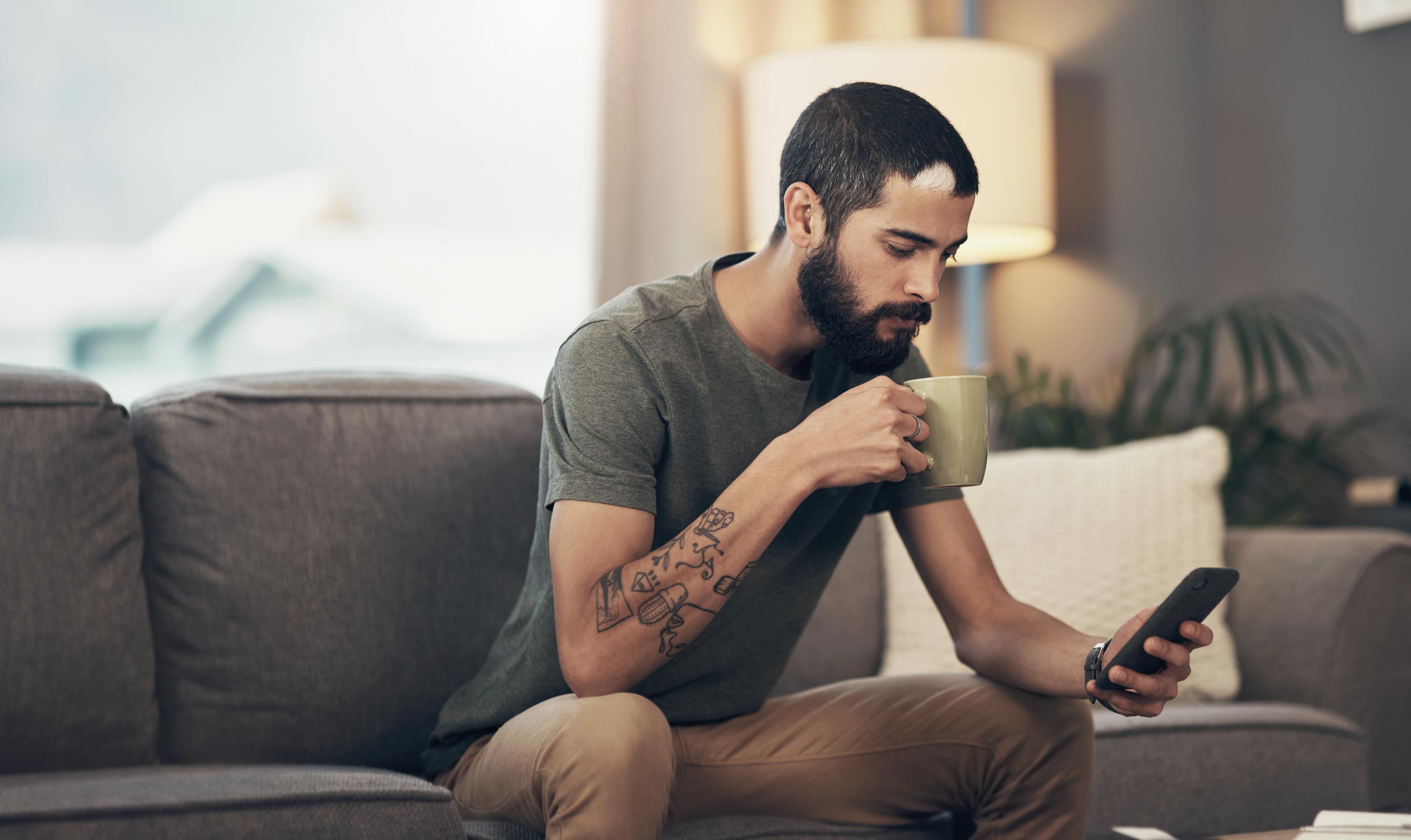 Slender young man with a beard, sitting on a couch drinking coffee and looking at the YOPX app on his phone.